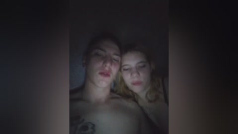 A video of two sleeping, nude women with fair skin and blonde hair. The image is dimly lit, creating a soft, intimate ambiance. The women's faces are close together, their eyes closed, and their expressions serene.