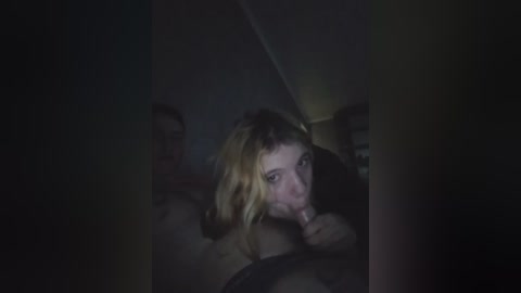 Video of a dimly lit scene with a young woman, possibly in her early 20s, with shoulder-length blonde hair, performing oral sex on an unseen man in a dark, confined space.
