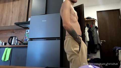 Video of a shirtless, tattooed man in beige shorts standing in a modern kitchen, with a fridge and wooden cabinets. Background shows a cluttered hallway and open door.