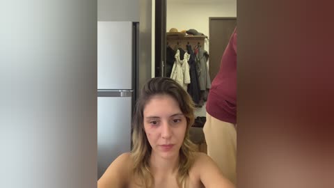 Video of a young, topless woman with light skin and wavy, light brown hair, looking into a mirror, showing a closet with hanging clothes in the background.