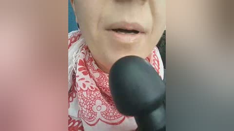 A close-up video of a woman wearing a red-patterned scarf, speaking into a black microphone. Her mouth is slightly open, and her light skin tone is visible. The background is blurred, with a focus on her face and the microphone.