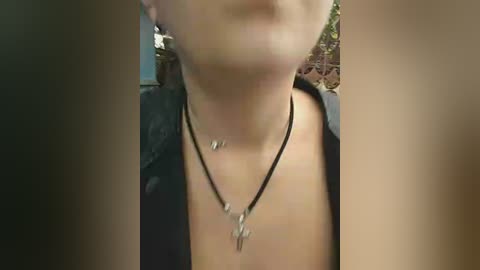 A close-up video of a woman with fair skin, wearing a black open shirt, black necklace with pendants, and a blurred background of greenery.