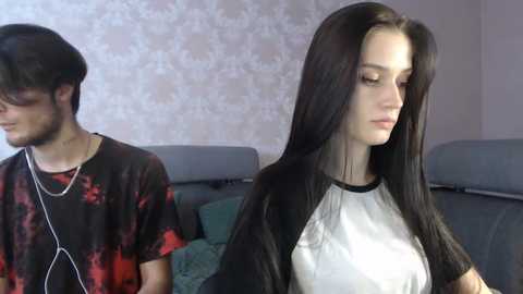 Media: Video of a young woman with long black hair in a white top, and a young man with dark hair and a red t-shirt, both sitting in a living room with patterned wallpaper and a gray couch.