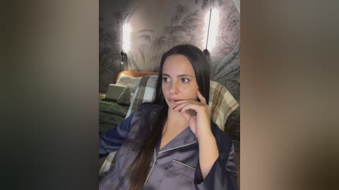 Media: Video of a woman with long black hair, wearing a dark blue satin robe, lying on a bed with green and beige pillows. The background features a mural and wall sconces.