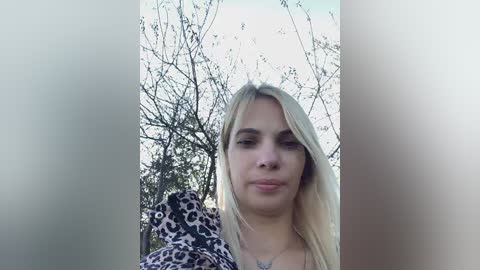 Media: A video of a blonde woman with straight hair, wearing a leopard print top, standing outdoors with a blurred tree and cloudy sky background.