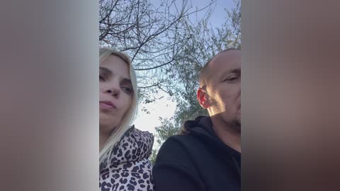 Media: Video of two people, a blonde woman with a leopard-print jacket and a bald man in a black hoodie, outdoors with bare trees in the background.