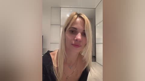 Media: Video of a blonde woman with straight hair, wearing a black top, standing in a modern, minimalist room with white walls and mirrors.