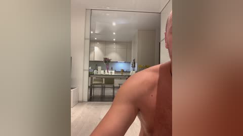 Media: Video of a shirtless, muscular man with light skin leaning against a white wall in a modern kitchen with beige countertops, stainless steel appliances, and a minimalist design.