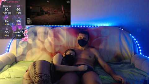 Video of two shirtless men, one Asian and the other white, wearing face masks, lying intimately on a colorful bed with blue LED lights.