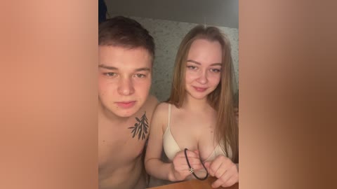 A video of a young, fair-skinned couple with medium build and light brown hair. The man, shirtless, has a tribal tattoo on his chest. The woman wears a white bra, smiling. They are indoors, possibly in a bedroom, with blurred background.
