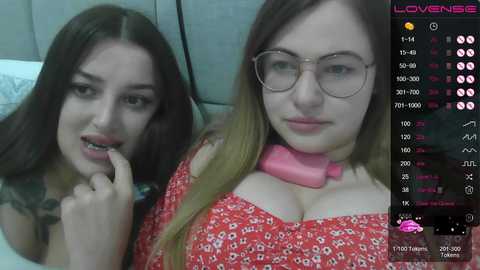 Video of two young women with pale skin, one with long dark hair, one with long blonde hair, both wearing glasses. They are in a bedroom, smiling.