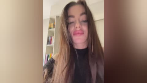 Video of a young woman with long, wavy brown hair, wearing a black turtleneck, smiling slightly, standing in a room with bookshelves in the background.