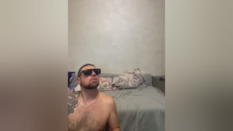 Video of a shirtless, tattooed man in dark sunglasses, standing in a dimly lit bedroom with a bed covered in gray sheets and a patterned pillow.