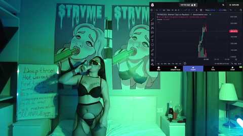 Video of a woman in black lingerie, eating a green lollipop, in a dimly lit bedroom with green lighting. A computer screen shows a graph and chat window.