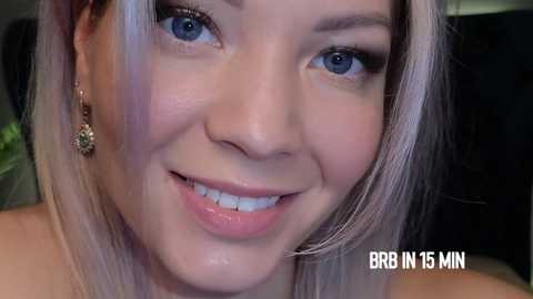 Media: Close-up video of a smiling Asian woman with fair skin, blue eyes, and light blonde hair, wearing gold earrings, in a dimly lit room. Text overlay reads \"BRR IN 15 MIN.\
