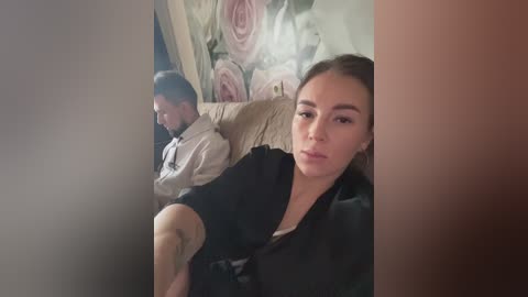 Media: Video of a woman with light skin, brown hair, and a small tattoo on her left arm, sitting on a beige couch in a dimly lit room with floral wallpaper.