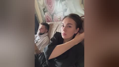 Media: Video of a young woman with long dark hair, wearing a black top, sitting next to a man with a beard, lying on a bed with floral wallpaper.