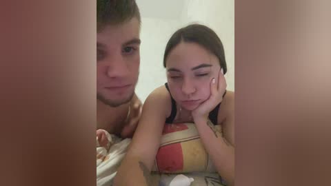 Video of a shirtless man with short brown hair and a young woman with long dark hair and a black top, both lying on a bed with a striped blanket, partially obscured by vertical lines.
