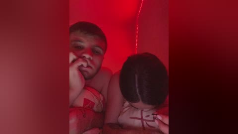 A video in red lighting shows a young, fair-skinned, bald man with a worried expression, lying next to a dark-haired woman in a white shirt. Both are in bed, creating a tense, intimate atmosphere.