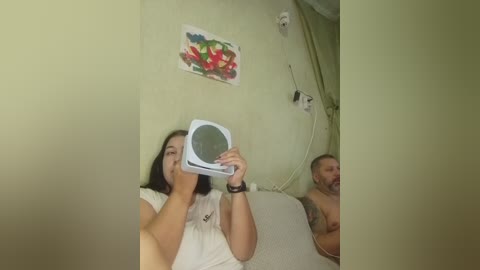 Video of a woman with dark hair, wearing a white top, taking a selfie with a white video camera. A man in a tank top is visible in the background, smiling.