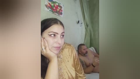 Video of a woman in a gold sari resting her head on her hand, looking pensive, while a shirtless man with a beard and tattoos lies on a bed behind her.