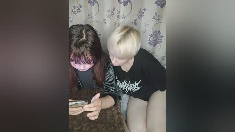 Media: A video shows a woman with long, dark hair and a man with short, blonde hair, both wearing black clothes, playing a video game on a carpeted floor. The background has a floral curtain.