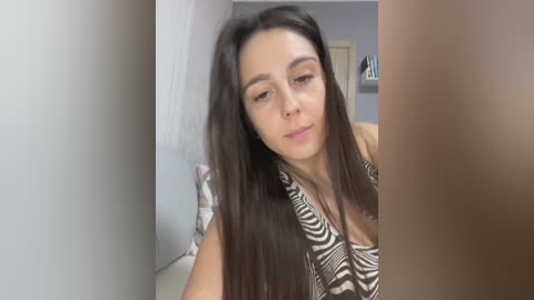 Video of a young woman with long dark hair, wearing a zebra-patterned top, standing in a narrow hallway with white walls, a closed door, and a floral-patterned cushion.