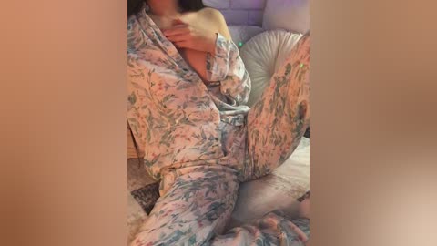 A video of a woman in a floral pajama set, seated on a bed with white pillows, her legs apart, revealing her genitals.