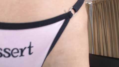 Video of a close-up of a woman's upper chest, showing part of a white top with black trim, revealing a small, silver ring piercing. Background includes beige curtains and a stack of papers.