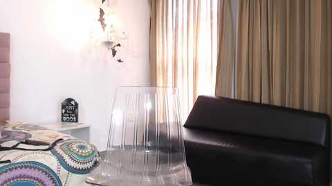 Video of a modern bedroom with a clear acrylic chair, beige curtains, a black leather sofa, and a bed with a colorful, mandala-patterned duvet.