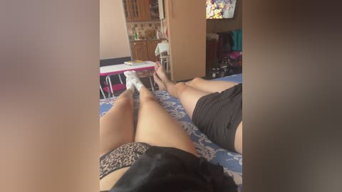 A video of a woman lying on her back on a blue-patterned bed, wearing leopard-print panties and black shorts. The room is cluttered with a table, chairs, and a TV in the background.