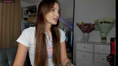 Video of a young woman with long brown hair in a white t-shirt, standing in a dimly lit room with shelves, a vase of flowers, and a camera in the background.
