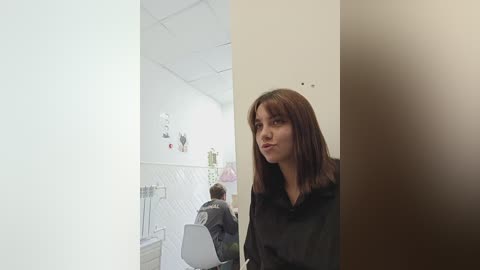 Media: A video of a young woman with long brown hair in a black robe, standing in a sterile, white-tiled hospital room with a medical bed and IV stands.