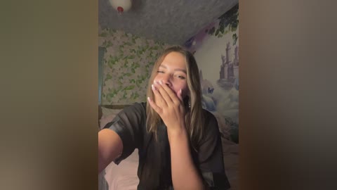 A video of a young woman with light skin and straight, shoulder-length brown hair, wearing a black shirt, covering her mouth with both hands, seated in a dimly lit bedroom with floral wallpaper and a bed.