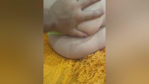 Video of a close-up, blurred hand squeezing a large, round breast, partially covered by a bright yellow bra. The subject is seated on a vibrant yellow shaggy rug.