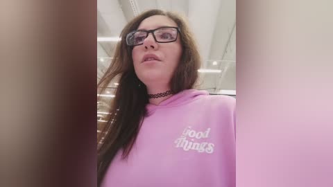 Video of a young woman with long brown hair, wearing black-framed glasses, a black choker, and a lavender sweatshirt with \"cool things\" in white text. She is indoors, with blurred people in the foreground.