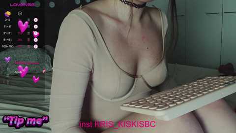 Video of a light-skinned woman with long brown hair, wearing a low-cut beige top and black choker, sitting at a keyboard, in a dimly lit bedroom.