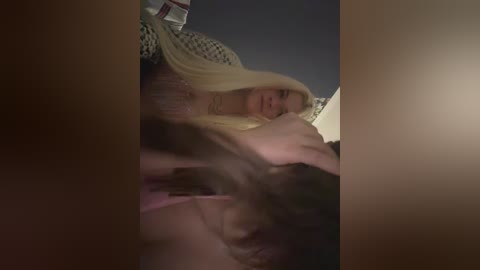 A video captures a blonde woman with fair skin, wearing pink lingerie, lying on a bed, partially obscured by a shadowy figure. The background features a dimly lit room with a white, patterned blanket.