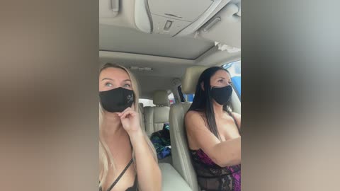 Media: Video of two women wearing black face masks, sitting in a car. The woman on the left has blonde hair, a black top, and a small breast size. The woman on the right has long black hair, a black top, and medium-sized breasts.