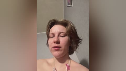 Media: A video of a fair-skinned, short-haired woman with closed eyes, wearing a pink necklace, standing in a beige-tiled bathroom with a framed picture on the wall.