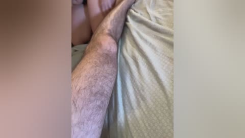 Video of a man's hairy, pale leg and arm lying on a bed with light gray, textured sheets.