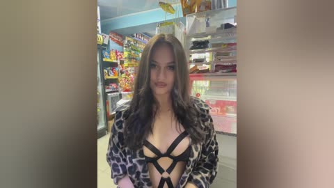 Media: Video of a confident woman with long brown hair, fair skin, and wearing a black, strappy leopard-print robe, standing in a brightly lit convenience store with shelves filled with colorful products.