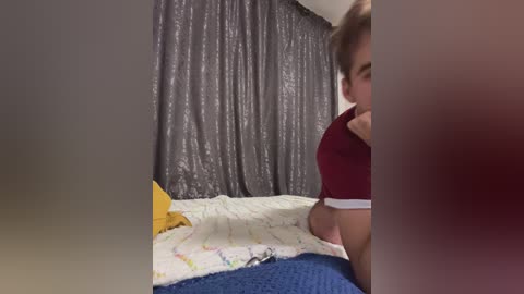 Video of a young woman with fair skin and brown hair in a red t-shirt, sitting on a bed with a blue blanket and yellow pillow, partially obscured by a grey curtain in a dimly lit room.