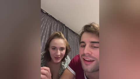 A video shows a young Caucasian couple with light skin, smiling and holding hands, in a dimly lit room with a gray curtain backdrop.