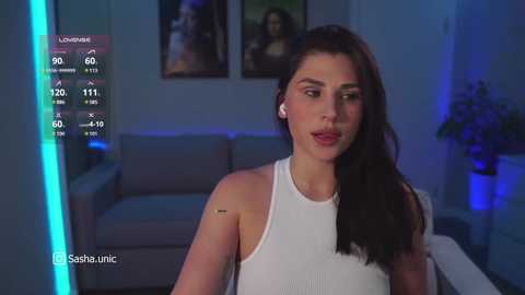 A video shows a Latina woman with long dark hair in a white sleeveless top, standing in a dimly lit modern living room. The background features a grey couch and a plant.