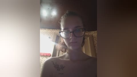 Video of a topless woman with glasses, light skin, and a tattoo on her shoulder, sitting in a dimly lit room with a polka-dotted blanket and a red pillow.