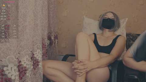 Video of a fair-skinned woman with short blonde hair, wearing a black face mask, black bra, and black panties, sitting on a chair in a room with floral-patterned wallpaper and a red and white lace curtain.