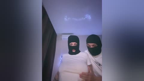 Video of two individuals, both wearing black balaclavas, standing indoors. The person on the left has a slight smile, while the person on the right has a neutral expression. The room has a dim, blue-toned lighting.