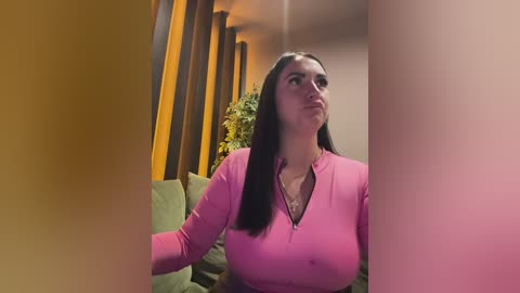 Video of a fair-skinned woman with long dark hair, wearing a pink zip-up top, sitting on a green couch in a dimly lit room with vertical yellow curtains and a potted plant.