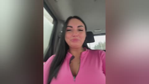 Video of a woman with long black hair and light skin, wearing a pink zip-up jacket, sitting in a car with a blurred window view.